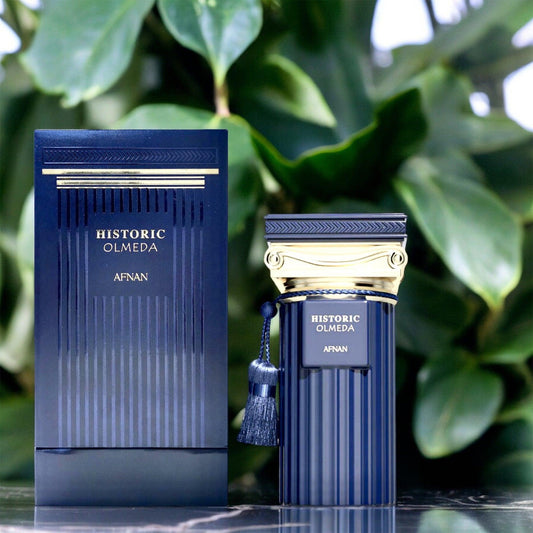 Afnan Historic Olmeda perfume bottle and box in deep navy blue with gold detailing, placed against lush green leaves, representing elegance and sophistication