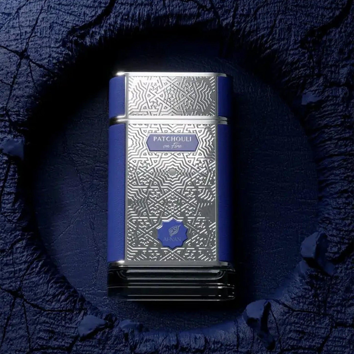 Afnan Patchouli On Fire Eau De Parfum bottle with intricate silver and blue design displayed on a textured dark blue surface with artistic lighting