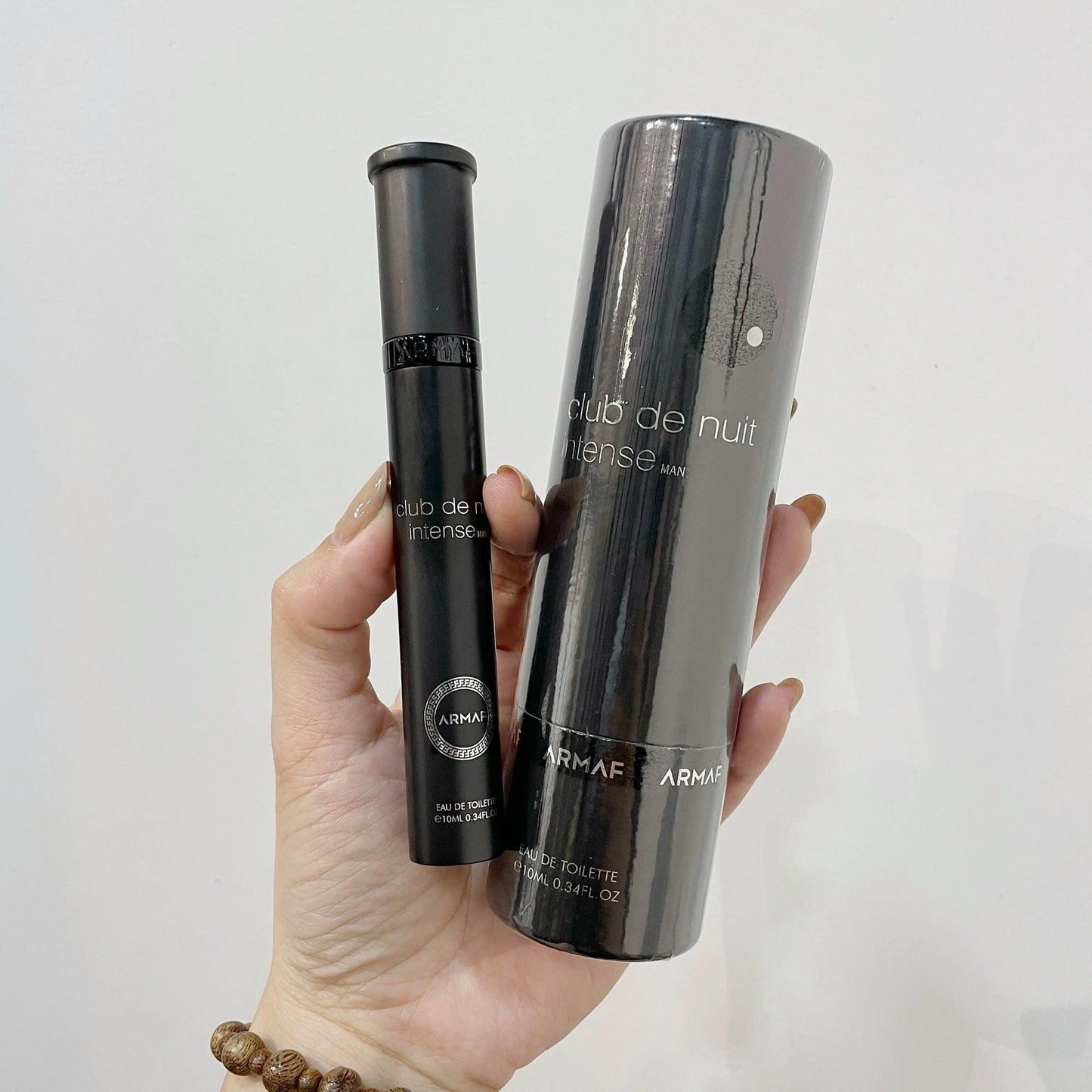 Hand holding Armaf Club De Nuit Intense Man travel perfume spray and cylindrical case in sleek black packaging, displayed against a light background for a clean, elegant look
