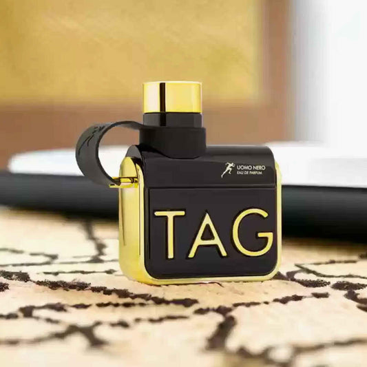 Armaf Him Tag Uomo Nero Eau De Parfum black and gold bottle placed on a patterned carpet with a blurred indoor background