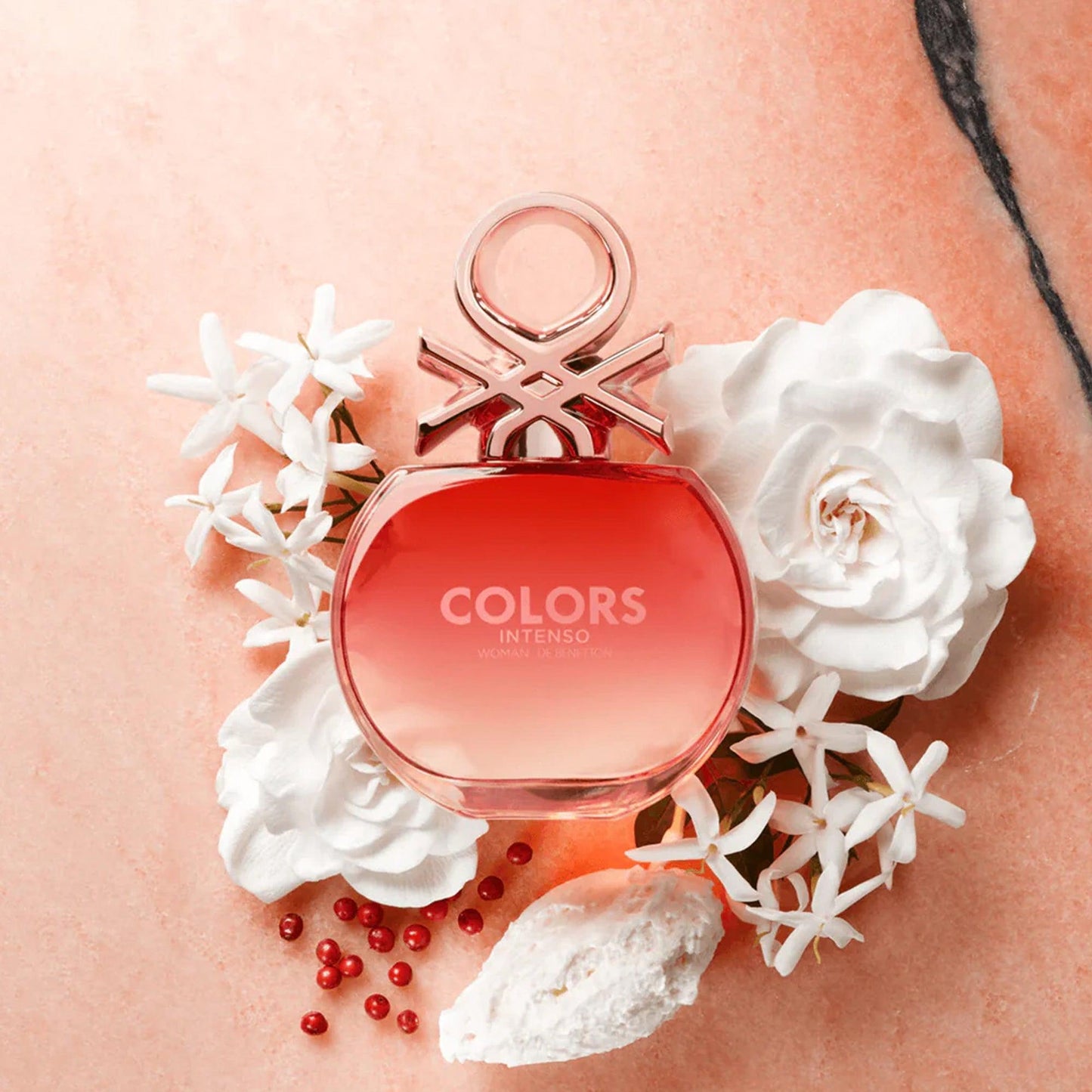 Benetton Colors Intenso Eau De Parfum pink gradient round bottle placed on a marble surface surrounded by white jasmine flowers, rose petals, and red berries