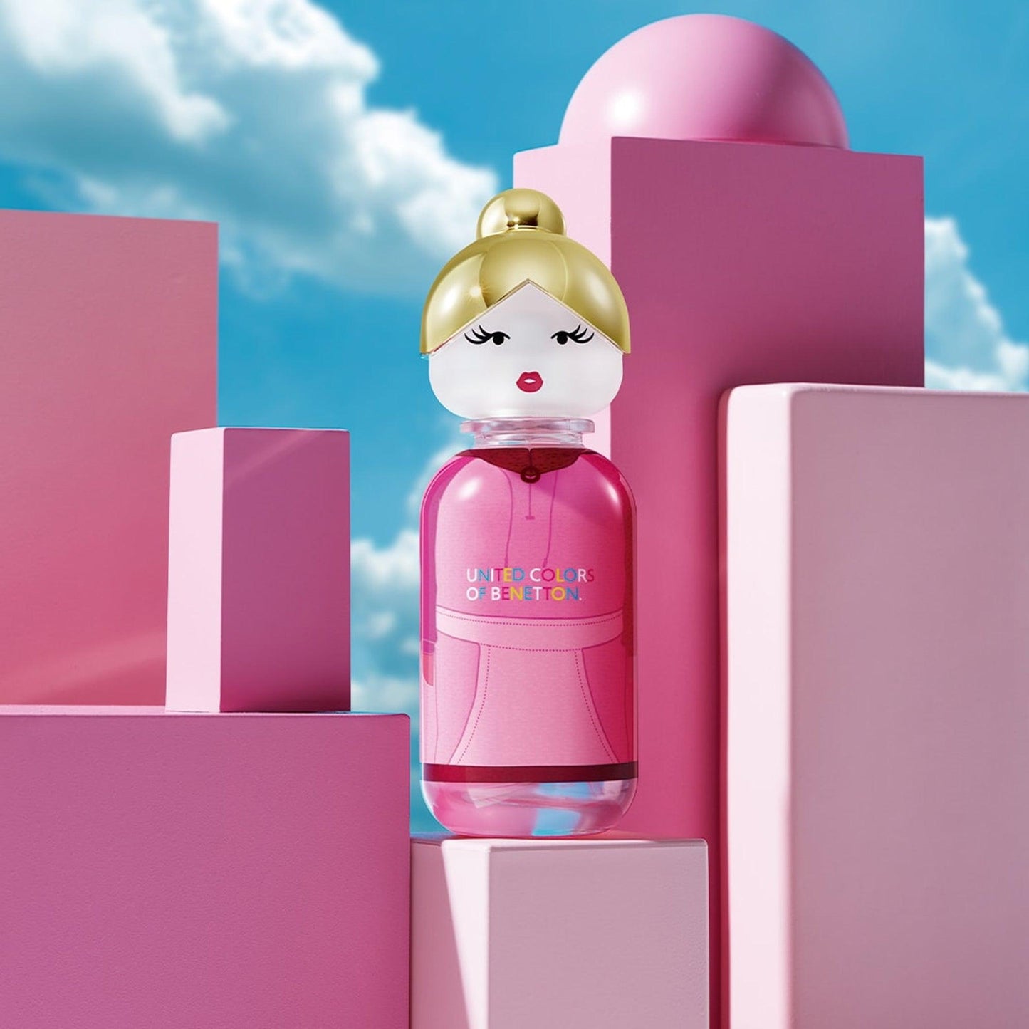 United Colors of Benetton perfume bottle designed as a pink doll with blonde hair and red lips, displayed among pink geometric blocks under a bright blue sky