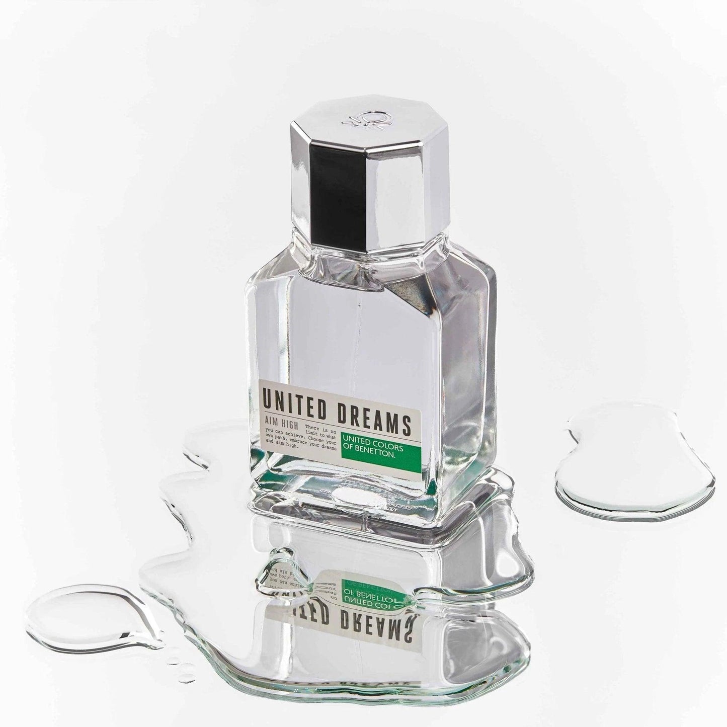 United Colors of Benetton United Dreams Aim High perfume bottle with a sleek silver cap, placed on a reflective surface with water ripples for a fresh, modern look