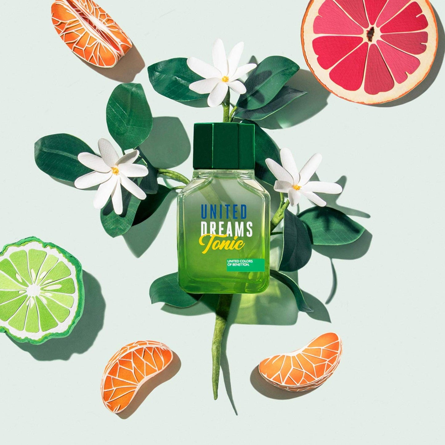 United Colors of Benetton United Dreams Tonic perfume bottle in bright green surrounded by citrus fruits, white flowers, and green leaves, reflecting a fresh and vibrant fragrance concept