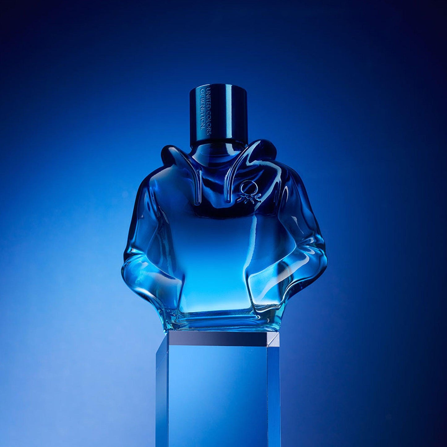 Jean Paul Gaultier Le Beau Le Male Eau De Toilette blue glass perfume bottle shaped like a hoodie displayed on a reflective pedestal with a dark blue gradient background