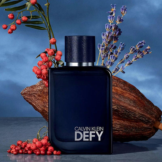 Calvin Klein Defy Eau De Parfum dark blue bottle surrounded by lavender, pink pepper, and woody elements on a moody blue background