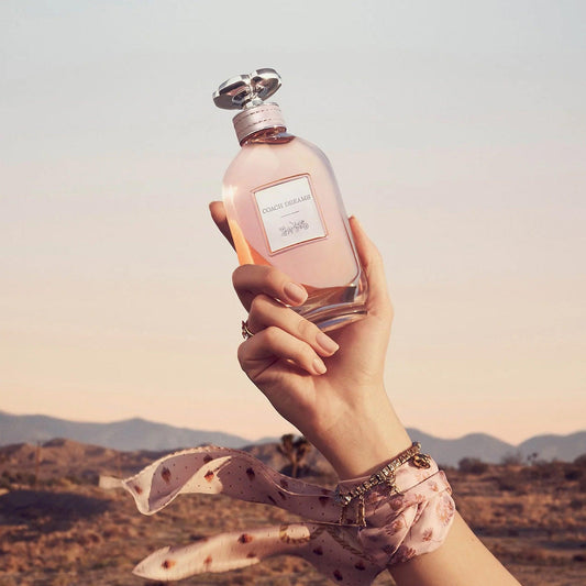 Hand holding Coach Dreams Eau De Parfum bottle against a scenic desert background during sunset, with a pink scarf wrapped around the wrist