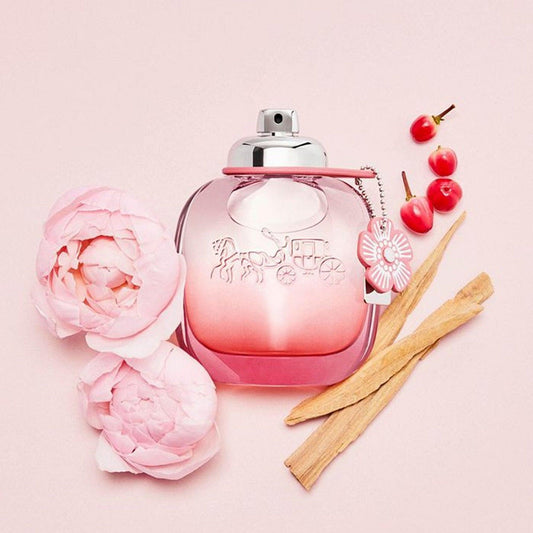 Coach Floral Eau De Parfum bottle in pink gradient glass, surrounded by soft pink peonies, cherries, and wood notes on a pastel pink background, evoking a fresh and feminine scent