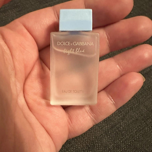 Hand holding a mini bottle of Dolce & Gabbana Light Blue Eau De Toilette with a frosted glass design and light blue cap against a dark background.