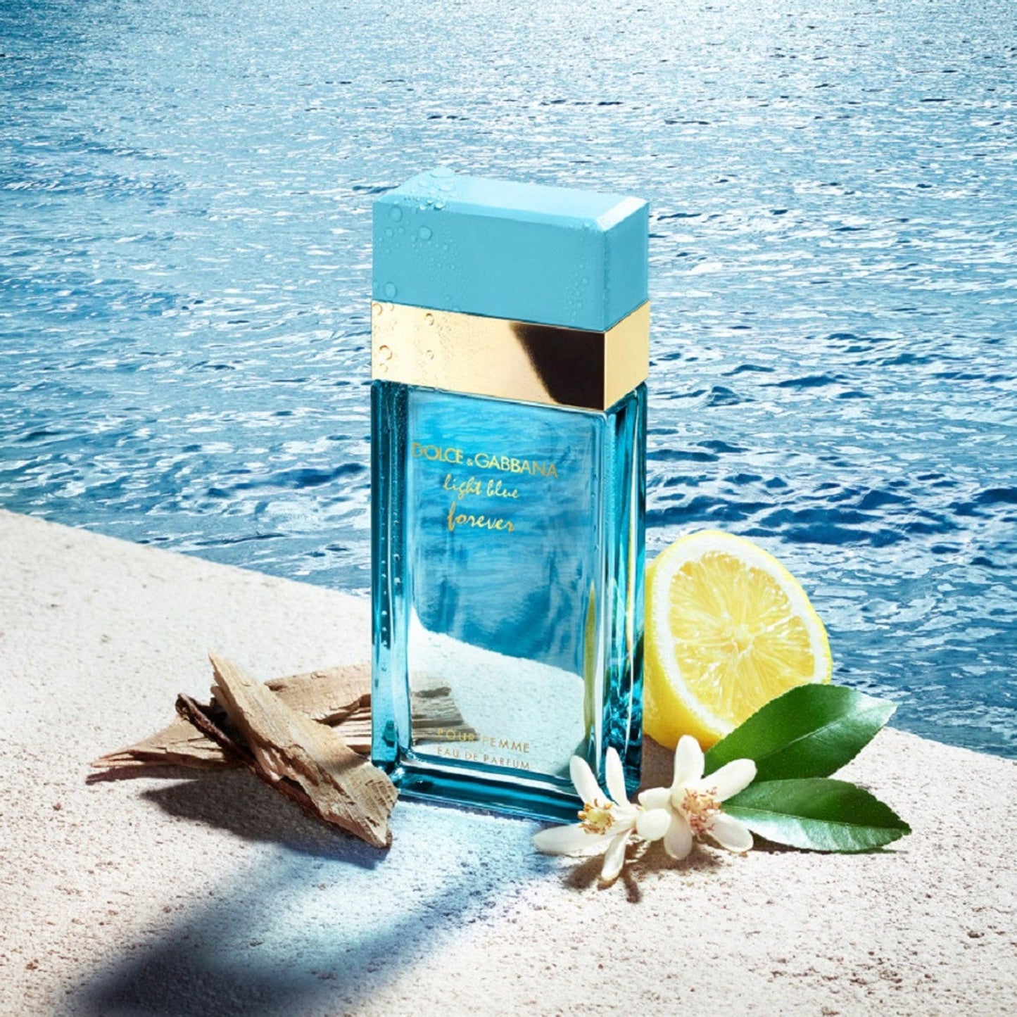 Dolce & Gabbana Light Blue Forever Eau De Parfum blue glass bottle placed near a lemon slice, white flowers, and driftwood beside a sparkling ocean background