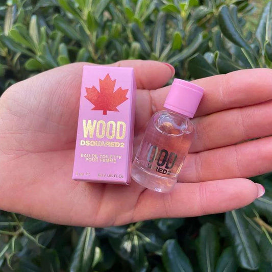 Hand holding a mini bottle and box of Dsquared2 Wood Pour Femme Eau De Toilette in pink packaging against a green leafy background.