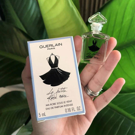 Hand holding Guerlain La Petite Robe Noire mini bottle and box against leafy green background