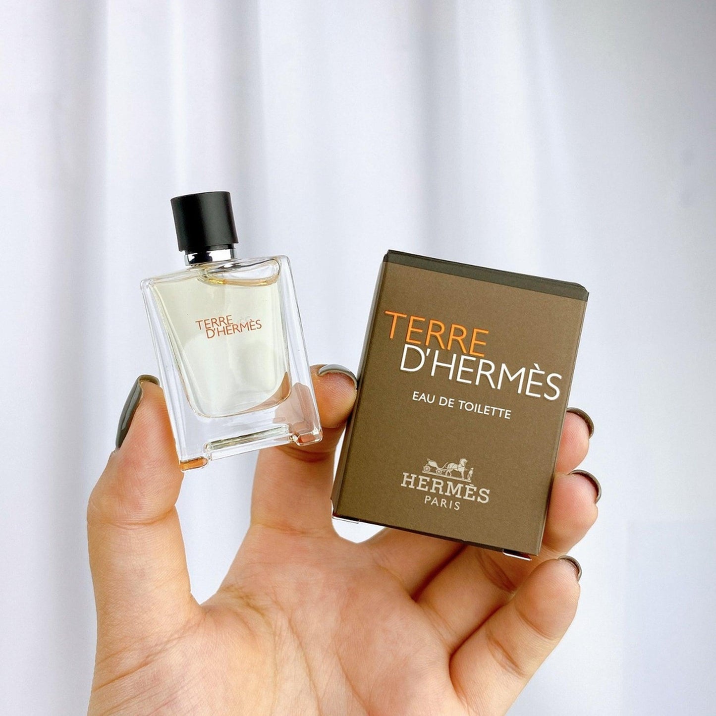 Hand holding a mini bottle and box of Terre d’Hermès Eau De Toilette by Hermès Paris against a white curtain background.