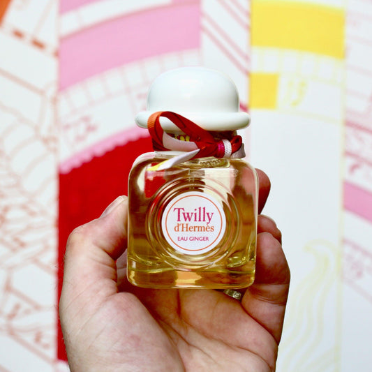 Hand holding a bottle of Hermès Twilly d’Hermès Eau Ginger perfume with a white cap and pink ribbon against a colorful abstract background.