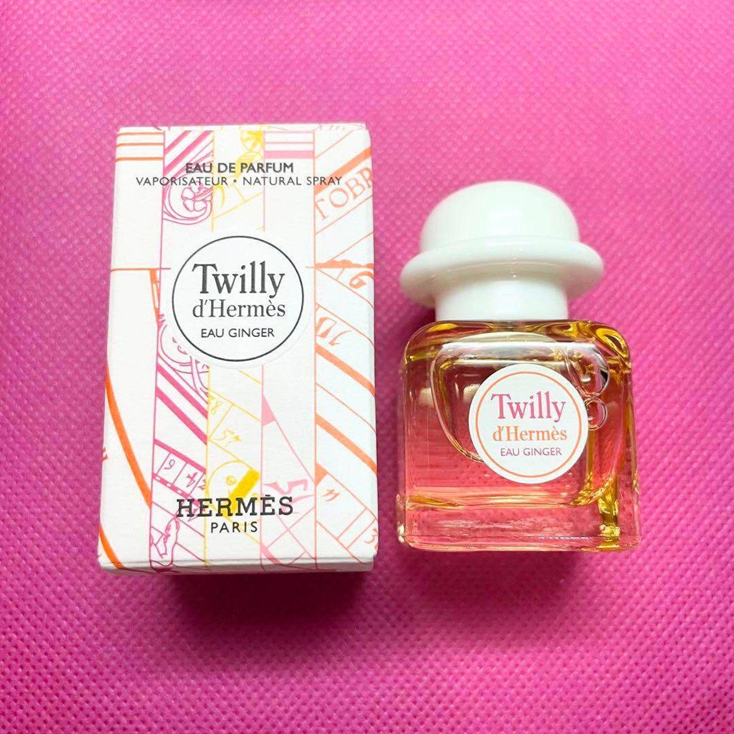 Hermès Twilly d’Hermès Eau Ginger Eau De Parfum bottle and box placed on a bright pink textured background.