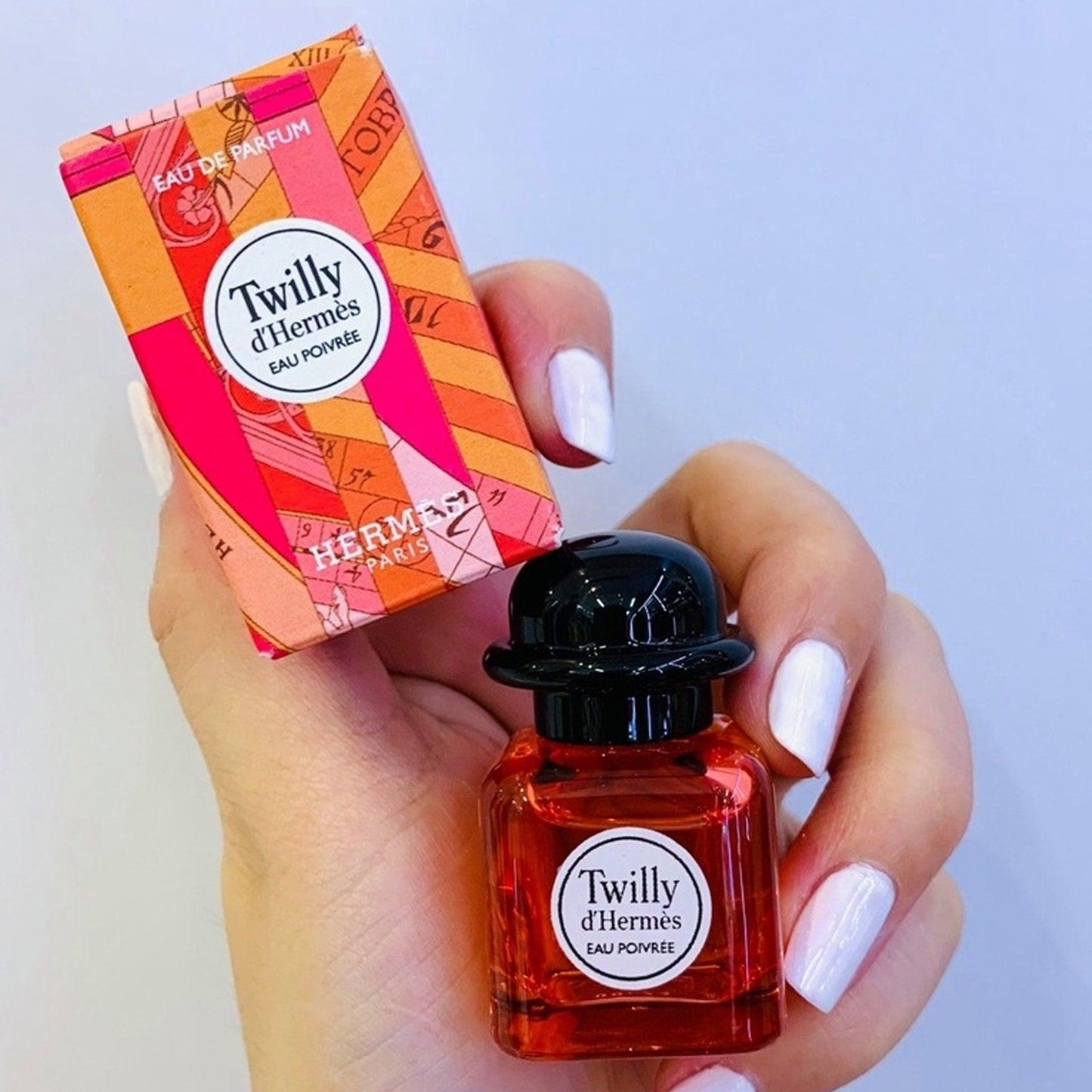 Hand holding Hermès Twilly d’Hermès Eau Poivrée Eau De Parfum mini bottle and matching pink-orange box against a light background.