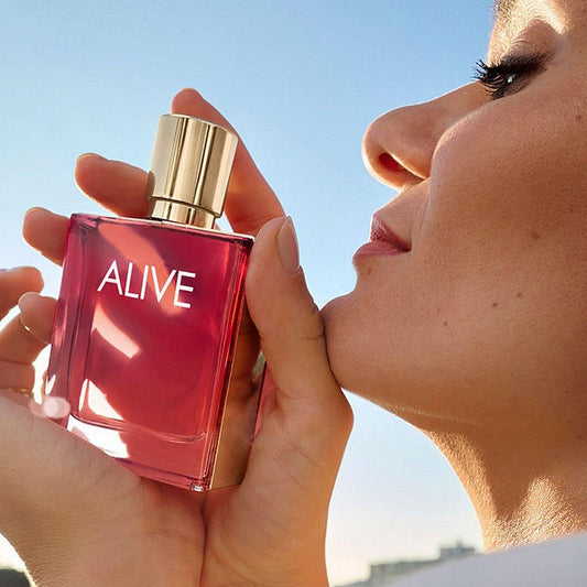 Close-up of a woman holding a red bottle of Hugo Boss Alive Eau De Parfum near her face under clear blue sky
