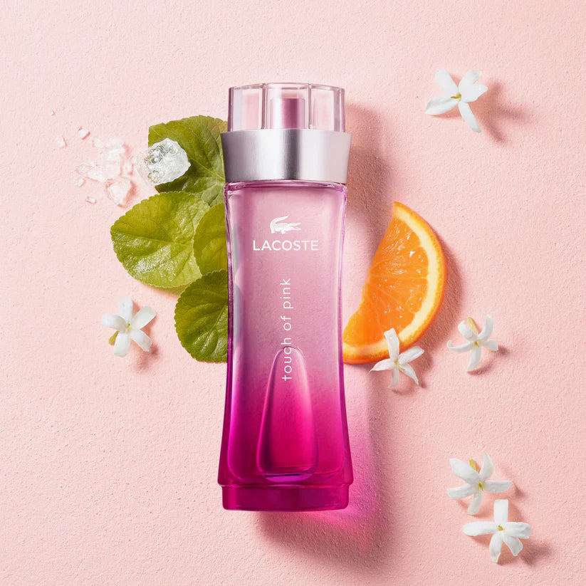 Lacoste Touch of Pink perfume bottle placed on a soft pink background with white flowers, green leaves, ice cubes, and an orange slice