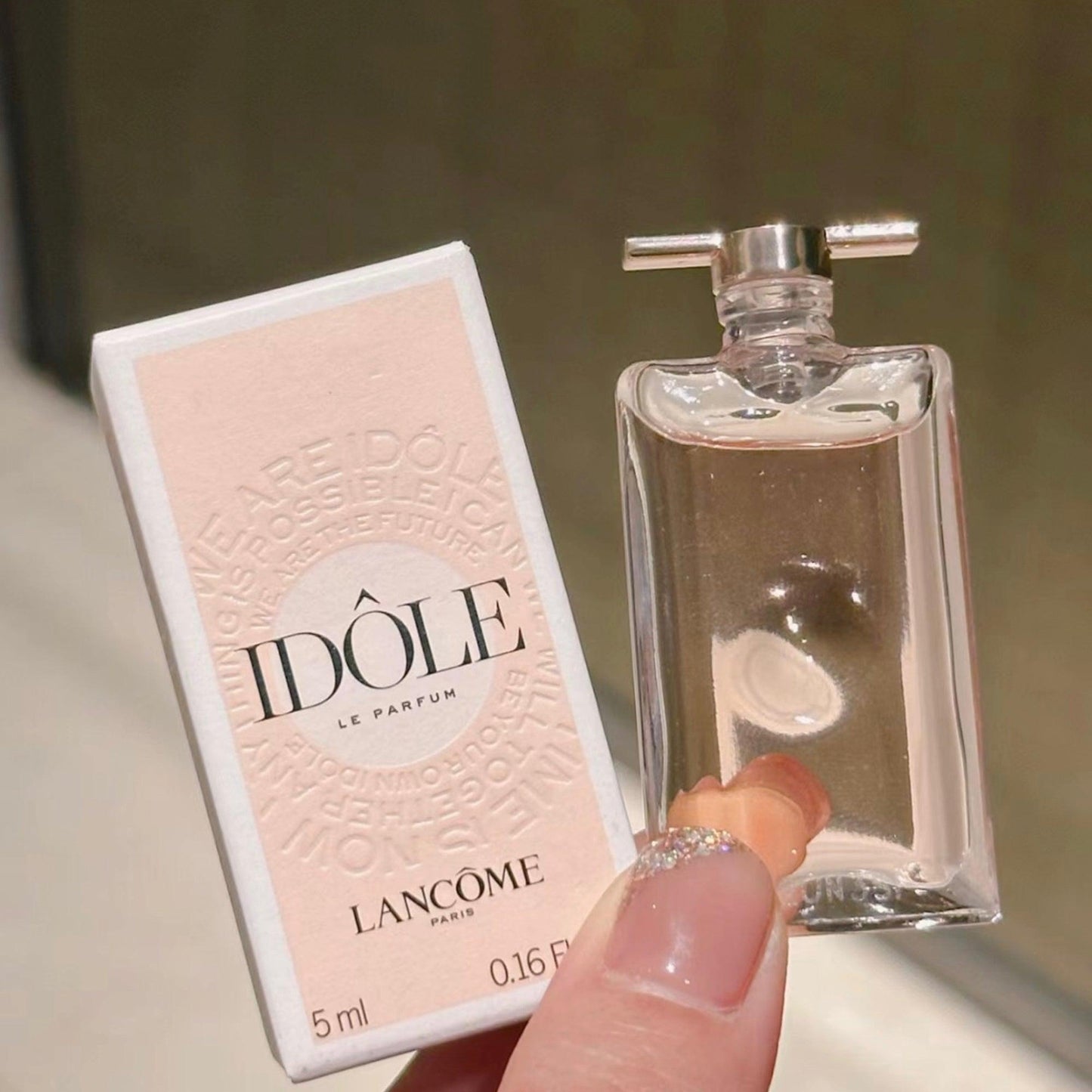 Lancôme Idôle Le Parfum 5ml bottle held by hand beside its pink and white packaging box with soft background