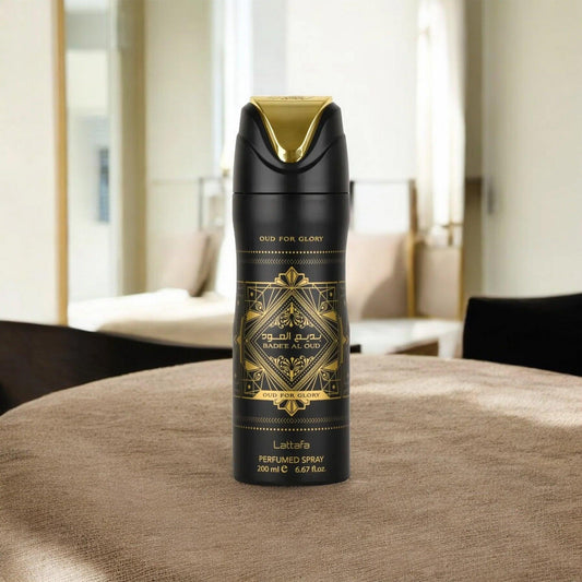 Lattafa Bade’e Al Oud Oud For Glory perfumed spray in a sleek black and gold can placed on a beige fabric surface with a blurred luxury room background, symbolizing elegance and opulence