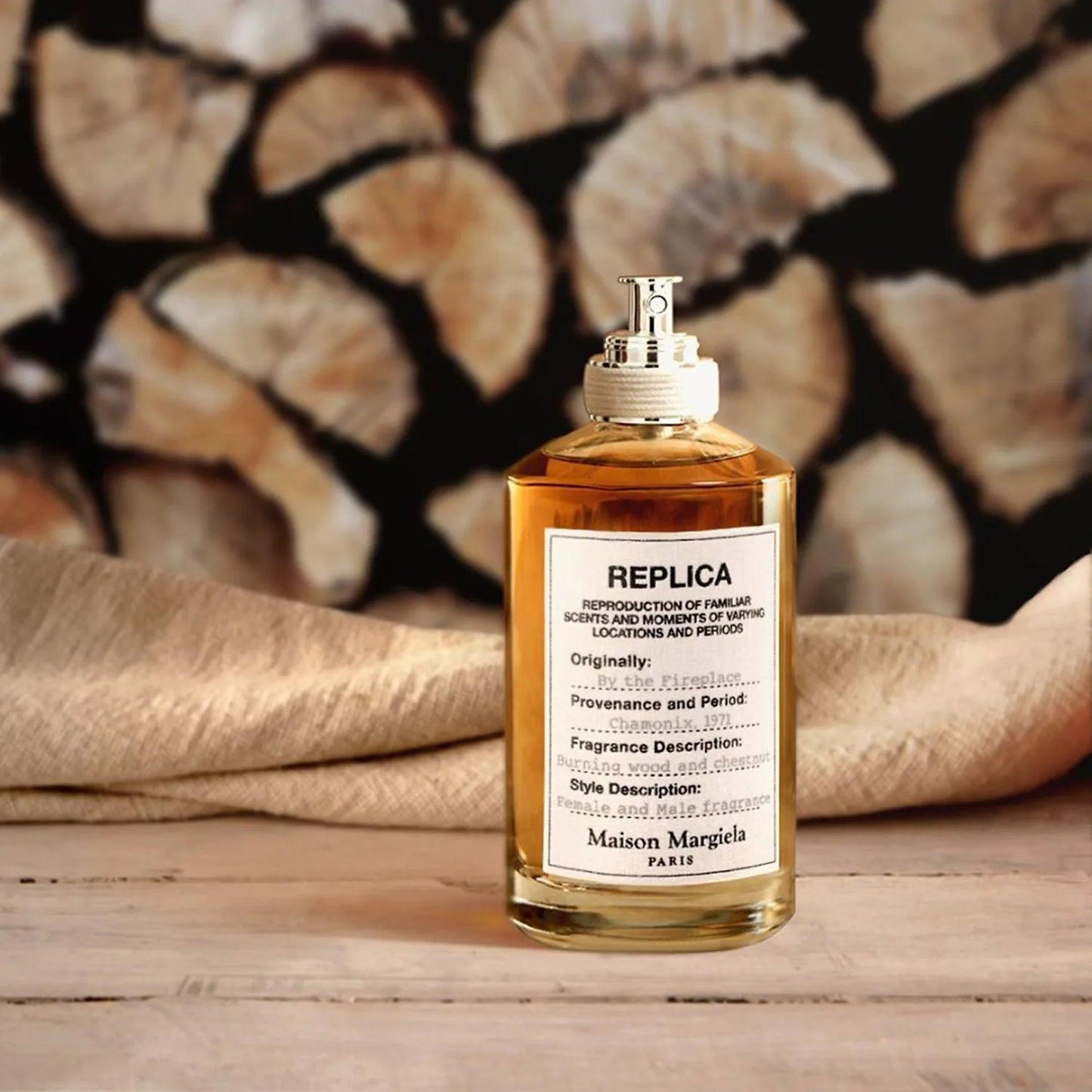 Maison Margiela Replica By the Fireplace Eau De Toilette bottle placed on a wooden surface with beige fabric and stacked firewood background