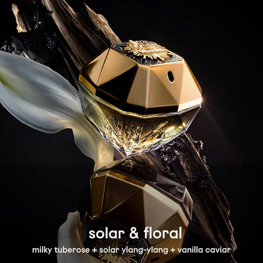 Paco Rabanne Lady Million Parfum diamond-shaped gold bottle placed on dark wood with white flower petals, reflecting a solar and floral theme with notes of tuberose, ylang-ylang, and vanilla