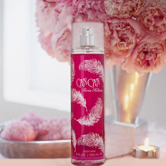 Paris Hilton Can Can Body Mist in a tall pink bottle with white feather design, placed beside soft pink flowers and a lit candle for a feminine, elegant look