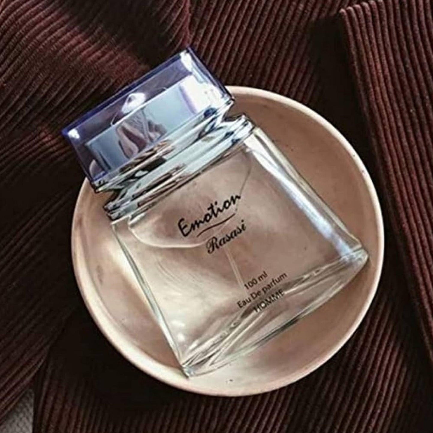 Rasasi Emotion Eau De Parfum 100ml bottle placed in a beige ceramic bowl on a dark brown textured fabric background.