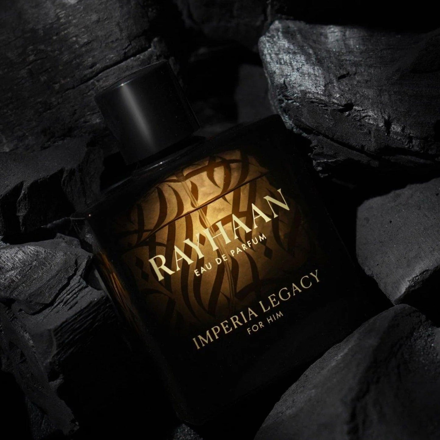 Rayhaan Imperia Legacy For Him Eau De Parfum bottle with golden glow surrounded by black charcoal rocks in a dark, moody background