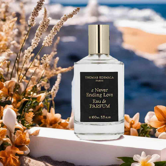 Thomas Kosmala A Never Ending Love Eau de Parfum bottle with gold cap placed outdoors among orange and white flowers overlooking a sunny seaside view