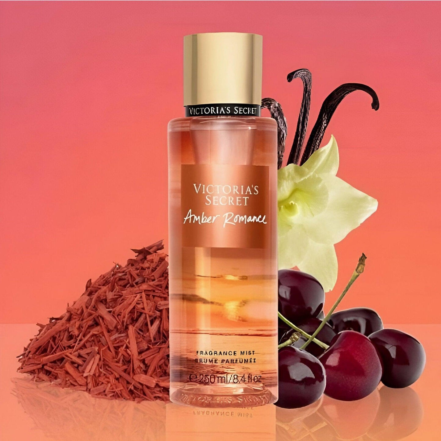 Victoria’s Secret Amber Romance fragrance mist bottle placed beside cherries, vanilla pods, and sandalwood on a glossy surface with a warm orange gradient background