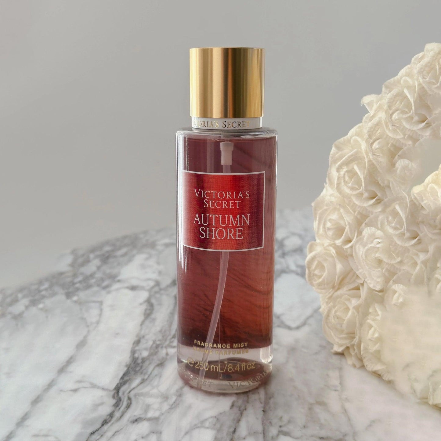 Victoria’s Secret Autumn Shore fragrance mist 250ml bottle with golden cap placed on marble surface beside white rose bouquet