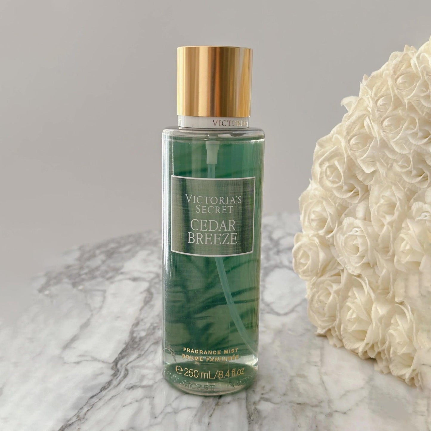 Victorias Secret Cedar Breeze fragrance mist 250ml bottle with gold cap placed on a marble surface beside white rose bouquet