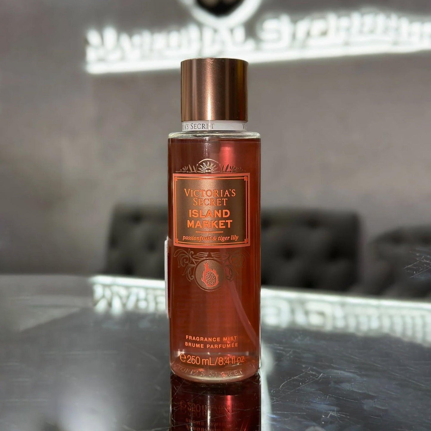 Victoria’s Secret Island Market fragrance mist 250ml bottle with bronze cap placed on reflective surface against a blurred grey background