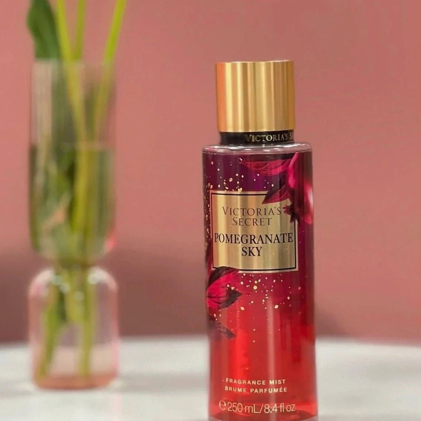 Victorias Secret Pomegranate Sky fragrance mist 250ml bottle with gold cap placed on a white surface against a soft pink background and vase with green leaves