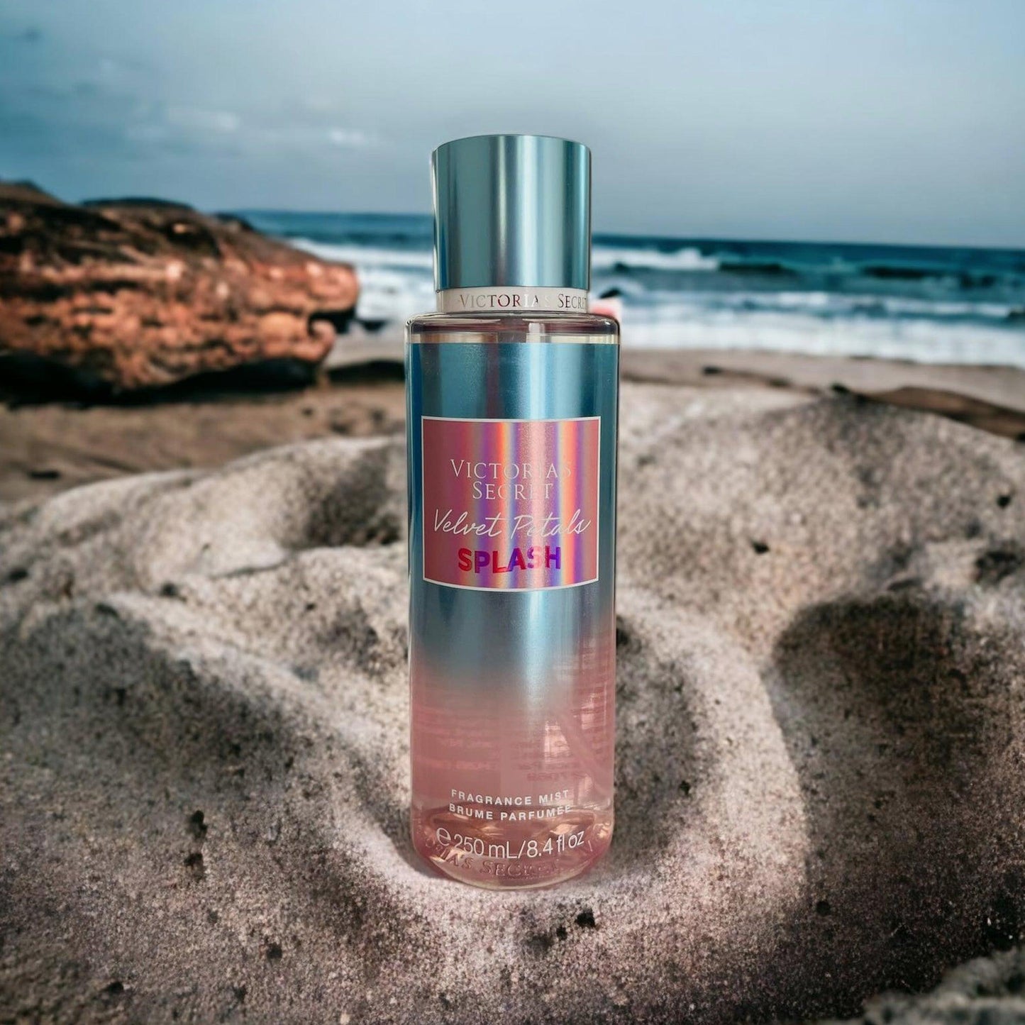 Victoria’s Secret Velvet Petals Splash fragrance mist 250ml bottle placed on sandy beach with ocean waves and rocks in the background
