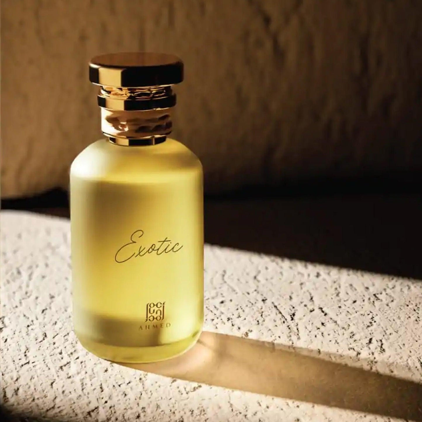 Rasasi Exotic Eau De Parfum 100ml bottle with gold cap placed on a textured beige surface under warm natural light and shadow