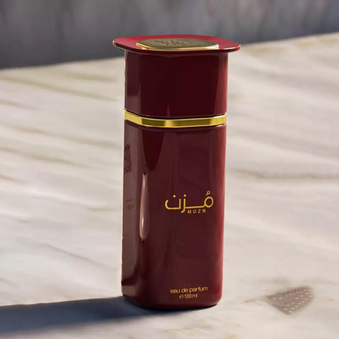Muzn Eau De Parfum 100ml bottle in a rich maroon color with gold detailing, elegantly placed on a marble surface under soft natural light
