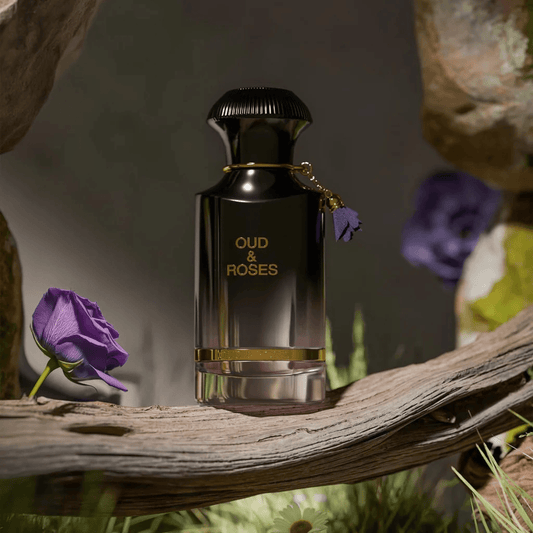 Oud & Roses Eau De Parfum bottle in glossy black and gold design placed on a wooden branch surrounded by purple roses and natural lighting