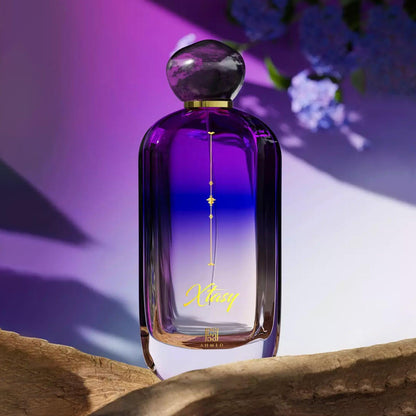 Ahmed Xtasy Eau De Parfum bottle in a striking purple-to-blue gradient design with gold detailing, placed on a wooden surface under soft purple lighting with flowers in the background