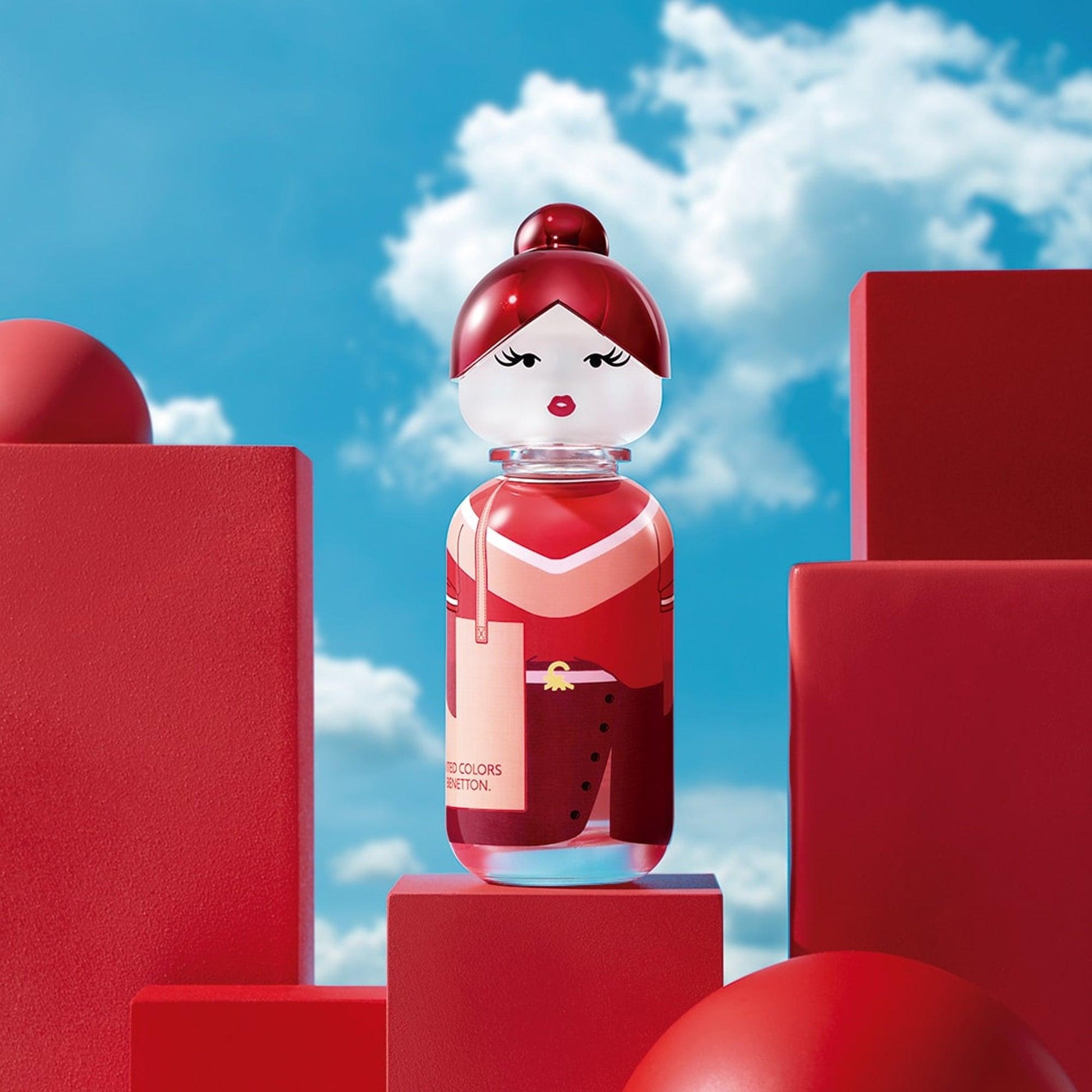 United Colors of Benetton red doll-shaped perfume bottle displayed among red geometric blocks with a bright blue sky background, symbolizing boldness and creativity