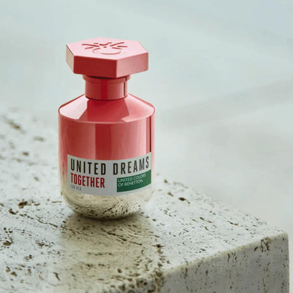 United Colors of Benetton United Dreams Together for Her perfume bottle in a pink metallic design, placed on a textured stone surface with natural lighting