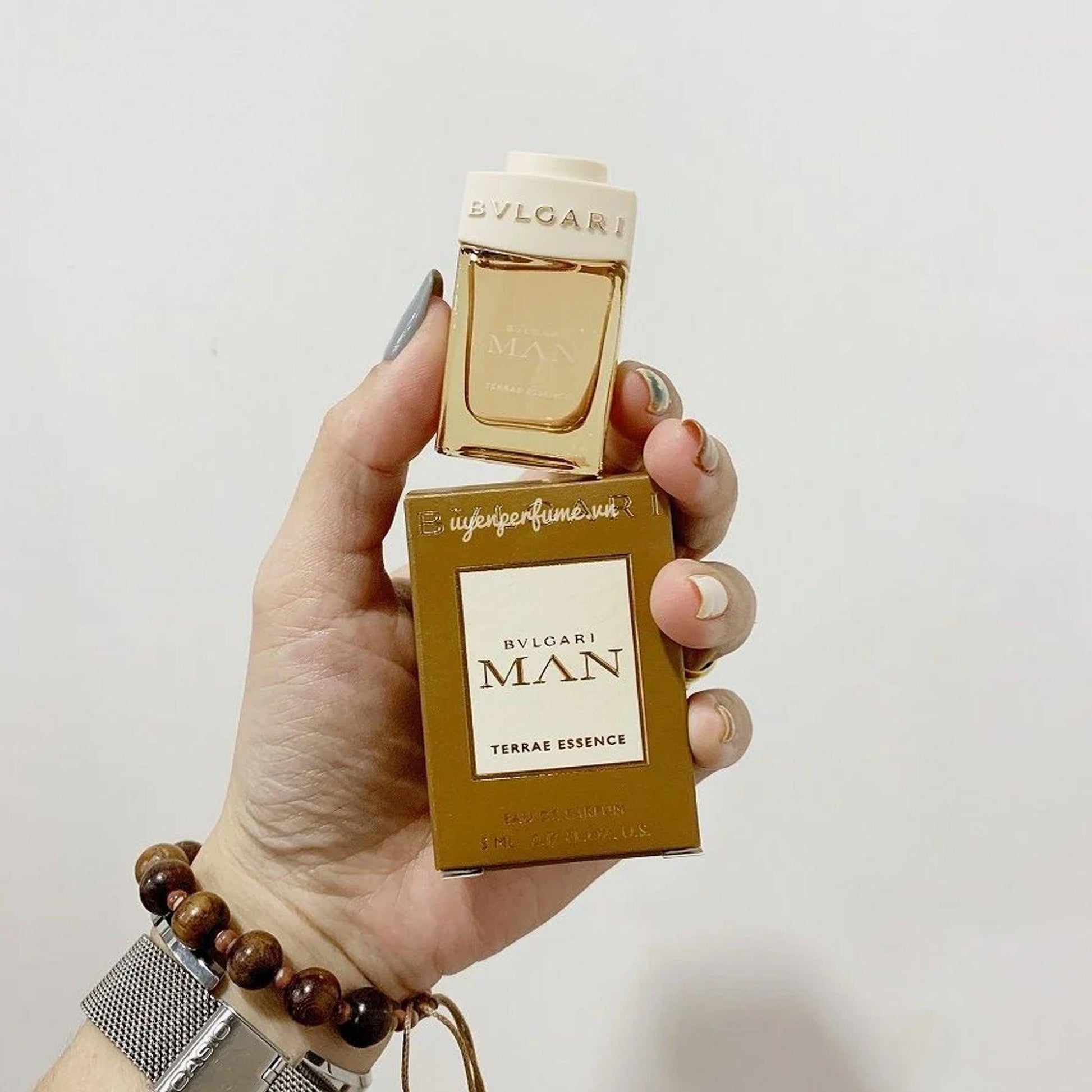 Hand holding a miniature bottle and box of Bvlgari Man Terrae Essence perfume, featuring elegant earthy tones and gold accents symbolizing sophistication and warmth