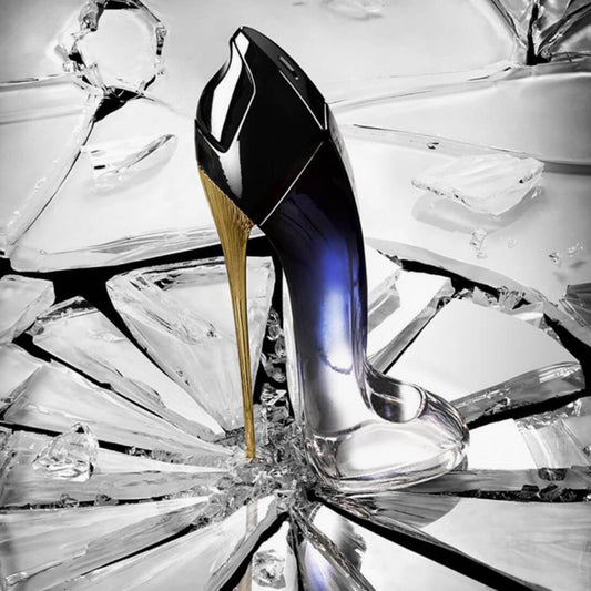 Carolina Herrera Good Girl perfume bottle shaped like a high-heeled shoe, featuring a gradient black-to-clear design with a gold stiletto heel, placed on shattered glass for a bold, glamorous effect