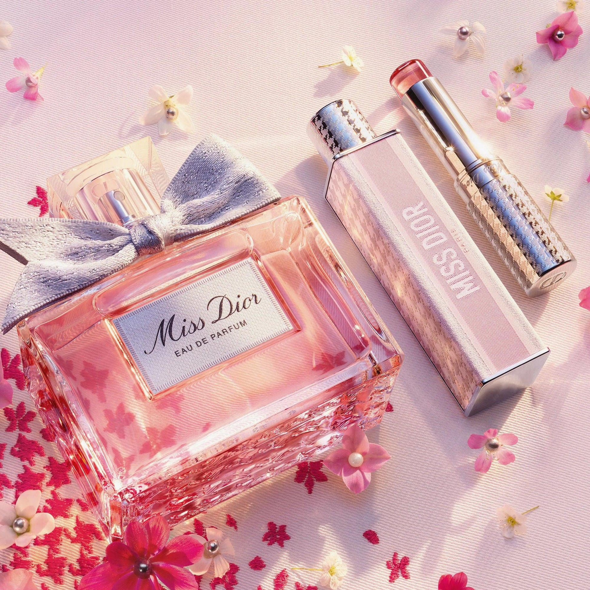 Miss Dior Eau De Parfum bottle and lipstick placed on a soft pink surface surrounded by small cherry blossom flowers, reflecting elegance and femininity