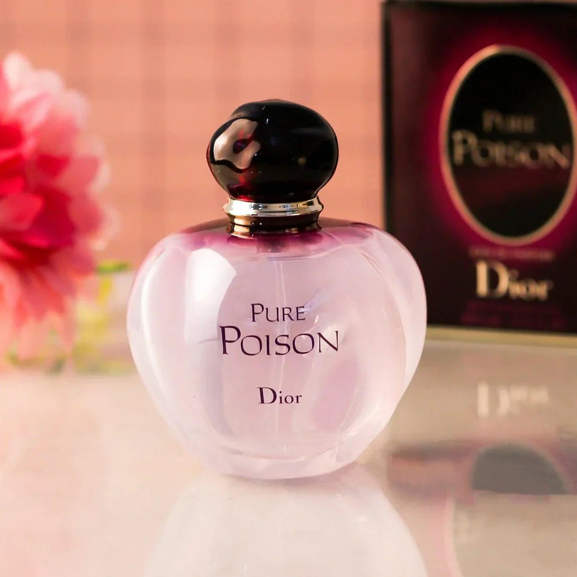 Dior Pure Poison Eau De Parfum bottle with a pearly white and purple gradient design placed on a reflective surface beside its packaging box and a pink flower in the background