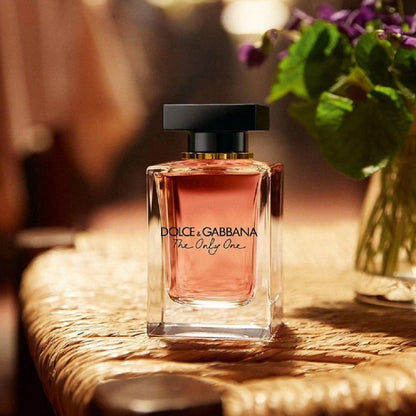 Dolce & Gabbana The Only One Eau De Parfum elegant glass bottle with amber-pink liquid placed on a woven surface beside a vase of flowers in warm indoor lighting