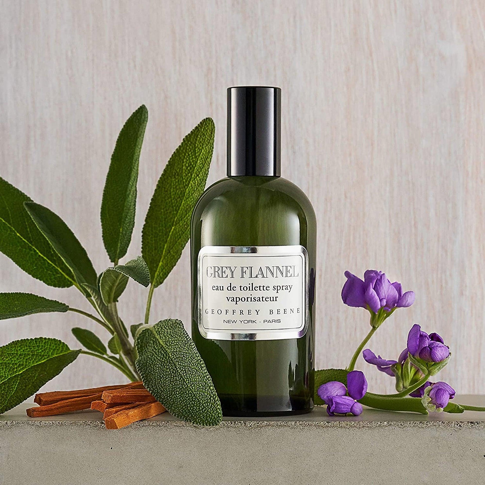 Geoffrey Beene Grey Flannel Eau De Toilette green bottle placed beside sage leaves, cinnamon sticks, and purple flowers on a neutral background