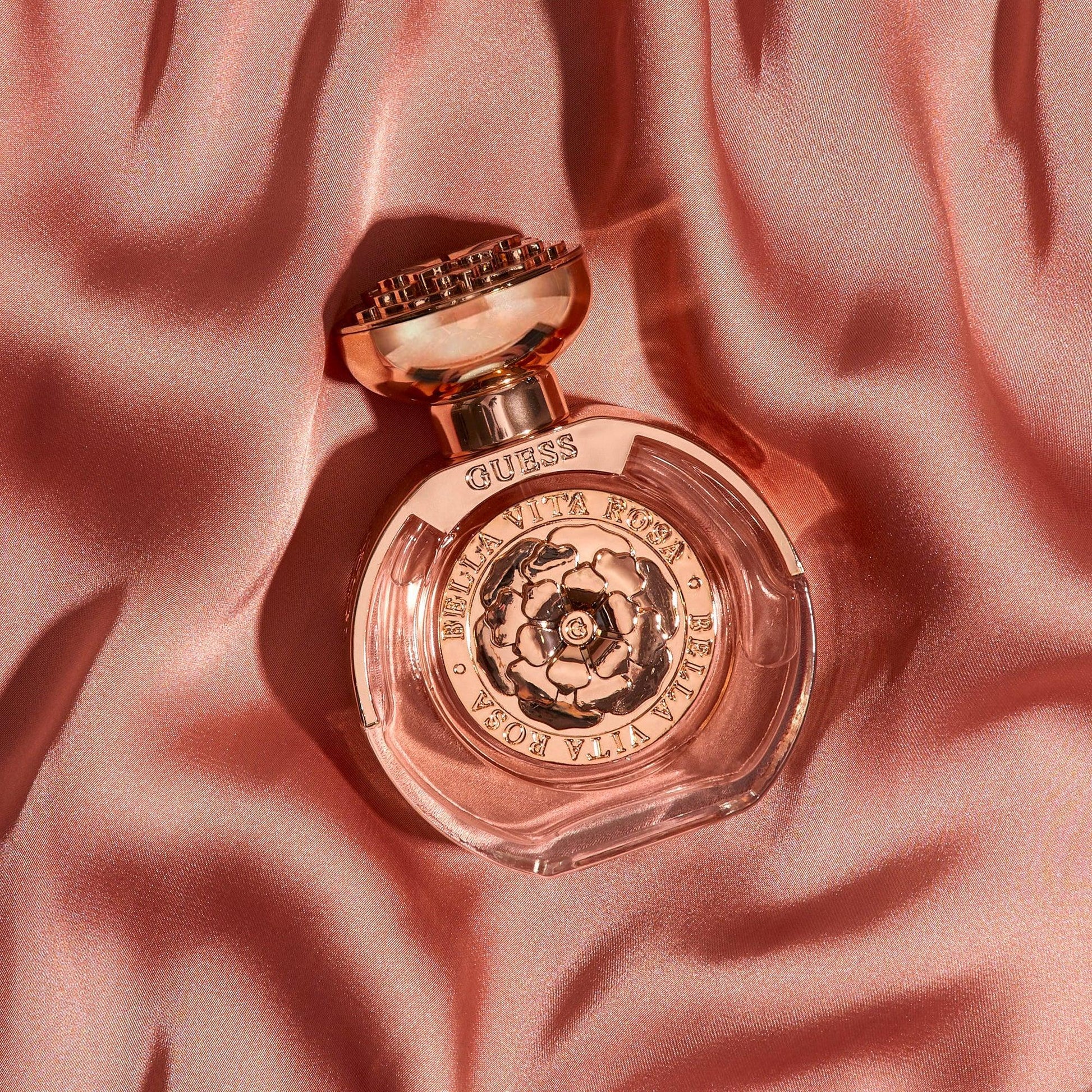 Guess Bella Vita Rosa perfume bottle with rose gold detailing placed on soft pink satin fabric under warm lighting