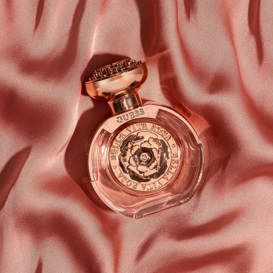 Guess Bella Vita Rosa perfume bottle with rose gold detailing placed on soft pink satin fabric under warm lighting